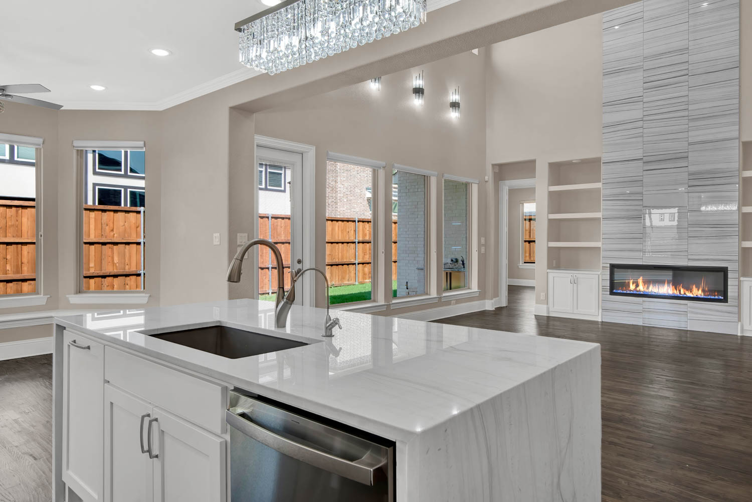 Modern kitchen with a large white island, double sink, and dishwasher. Spacious living area features floor-to-ceiling windows, a sleek fireplace, and built-in shelves.